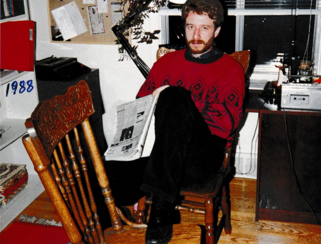 Man sitting on a chair with his foot up on another chair, with typewriter, desk, and casette player in background